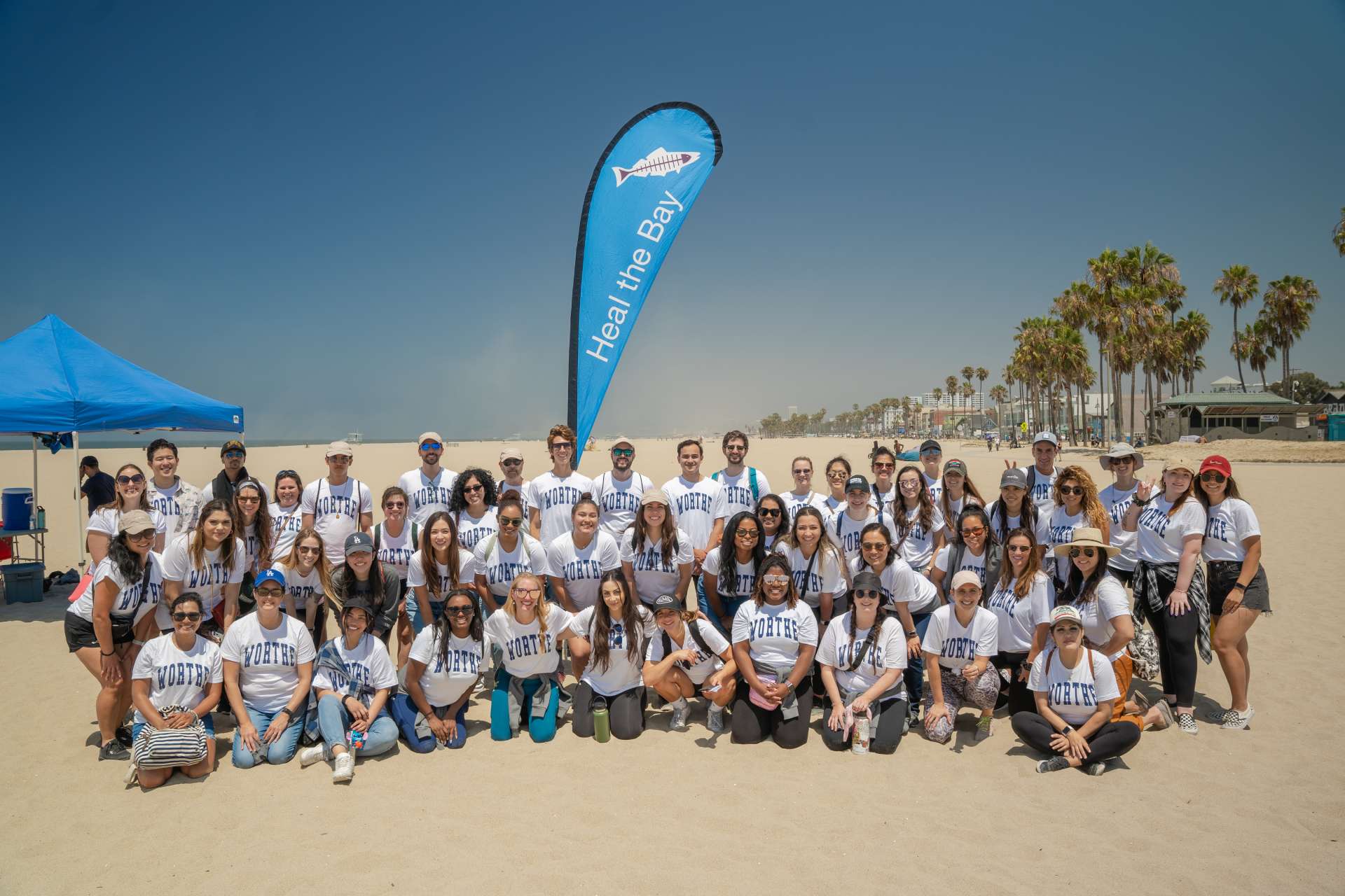 A group of about 50 people wearing matching shirts that bear the logotype 'Worthe' pose on a beach under a sign that says 'Heal the Bay'